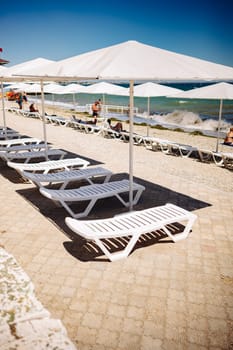 White sunbeds and beach umbrellas standing on the beach in Crimea during a sunny summer day, tourists relaxing and swimming in the sea