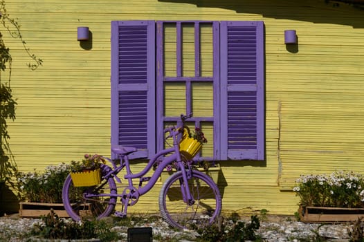 A delightful scene unfolds with a vivid purple bicycle, embellished with yellow flower planters, resting beside a sunny yellow wall. The colors bring joy and life to the surroundings.