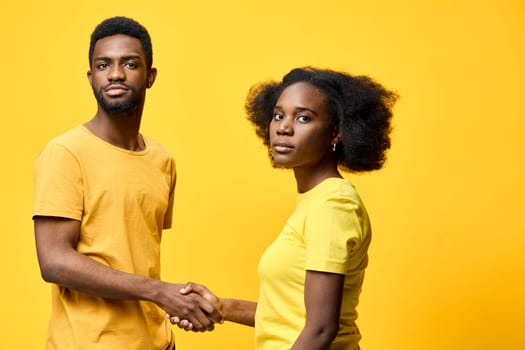 Two diverse young adults, a man and a woman in yellow shirts, engaging in a friendly handshake against a bright yellow backdrop, conveying unity and collaboration