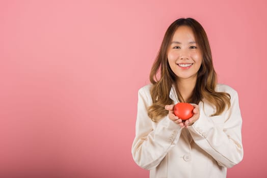 Smiling woman confidence showing holding red heart with her hand palm isolated pink background, Asian happy portrait beautiful young female send love and happy valentine in studio shot, copy space