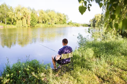 A man enjoys a peaceful day fishing by a serene lake, sitting in a comfortable chair, surrounded by lush greenery.