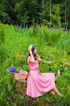 A young woman in a pink dress sits on a tree stump in a sunny forest glade, enjoying a picnic with wildflowers.
