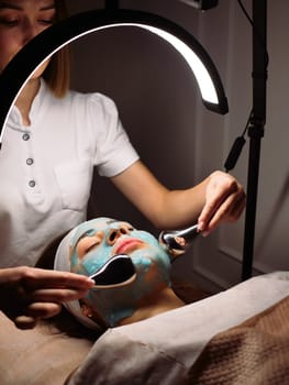 Cosmetologist does cryo massage with globules on blue hydrogel alginate mask in the beauty salon, vertical, close-up. Woman Facial Skin and Body Care