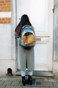 Portrait of a young Caucasian brunette girl from the back in full growth in a gray coat with a transparent backpack with a red calm sleeping cat, which sits inside and opens the front door with a key, returning from a walk, side view close-up. Concept of a visit to the vet, walks with pets.