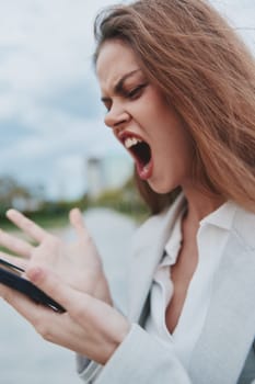 Frustrated businesswoman expressing anger towards her smartphone in an outdoor urban setting. Dressed in a stylish blazer, she embodies modern professionalism.