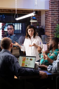 Diverse coworkers in startup office engaged in virtual meeting as caucasian woman leads the discussion. A large monitor displays video call with remote male colleague, highlighting modern teamwork.