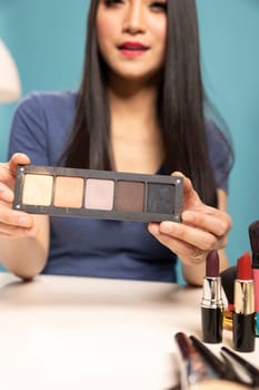 Young asian woman holding a cosmetic product up close, presenting it to the camera during her beauty vlog. Female beauty influencer showcasing an eyeshadow palette while recording for her channel.