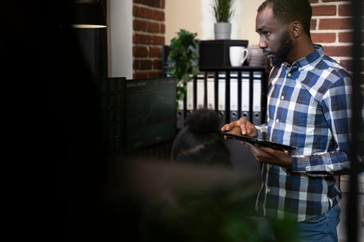 Male stock trader working late with his coworker, tracking financial market trends in a professional office setting. Selective focus of african american man using tablet during a meeting.
