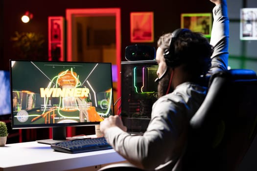 Happy pro gamer at home competing in intense multiplayer videogame, throwing fist in air after winning. Joyous person achieving victory in gaming session, overflowing with joy