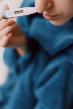 Woman holding electronic thermometer, eyes closed, wrapped in blue blanket.