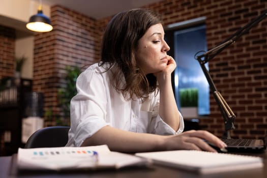 Caucasian female manager working tirelessly at her desk, leaning on one hand while scrolling through her laptop. A fatigued business analyst reviews company research data with an exhausted expression.