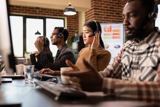 Multiethnic call center agents work at their stations, engaging with remote customers and processing service requests. Computers, coffee cups, and headsets set the scene for a busy office.