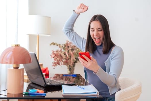 Excited Caucasian woman sitting at home office desk, looking at smartphone with joy. Laptop, notebook, and decor in modern workspace. Happy reaction, technology, communication, and digital interaction