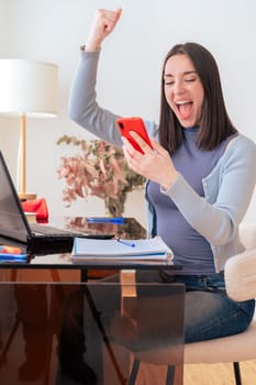 Excited Caucasian woman sitting at home office desk, looking at smartphone with joy. Laptop, notebook, and decor in modern workspace. Happy reaction, technology, communication, and digital interaction