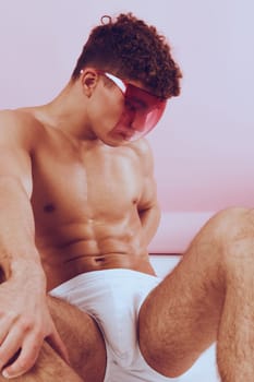 Fashion-forward young man with curly hair poses in pink sunglasses and white briefs on a soft, pastel background, showcasing modern style and confidence.