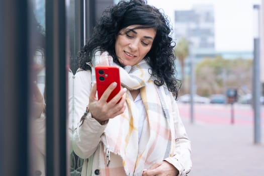 Happy caucasian woman standing outdoors in urban setting, wearing coat and scarf, using her red smartphone. She is smiling and relaxed, enjoying a casual moment on a cold day in the city.