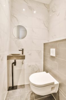 a white toilet in a small bathroom with marble tiles on the walls, and a round mirror hanging above it