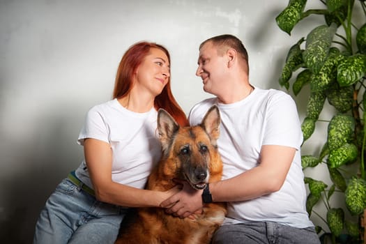 A couple sits closely together holding their beloved German Shepherd in a cozy room filled with indoor plants.