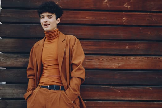 A fashionable young man wearing a brown turtleneck and suit jacket stands casually against a wooden backdrop The colors are warm, emphasizing his outfit