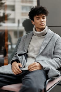 Fashion portrait of a young man sitting in front of a window wearing a grey sweater, black jacket, and white turtleneck.