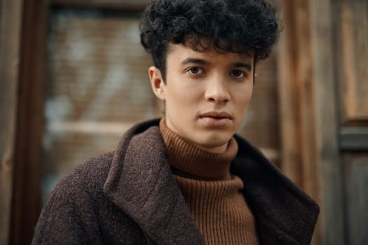 Fashionable young man with curly hair wearing a brown coat and a turtleneck sweater, standing in front of a wooden door and confidently looking at the camera.