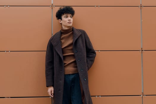 Fashionable young man in a brown coat and turtleneck sweater, standing casually with hands in pockets against an orange wall.