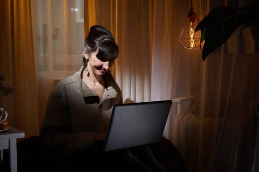 In a cozy apartment, a middle-aged woman finds pleasure in her evening routine while working on her laptop in soft light.
