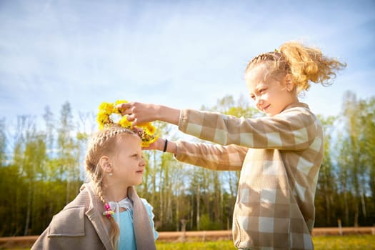 Two girls enjoy a sunny spring day, as one decorates the other with a flower crown in a vibrant green field.
