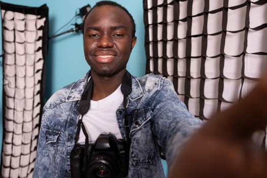 African american man records himself in a studio with photography equipment, using a camera. POV of black male photographer with a DSLR around his neck, smiling at the digital device in his hand.