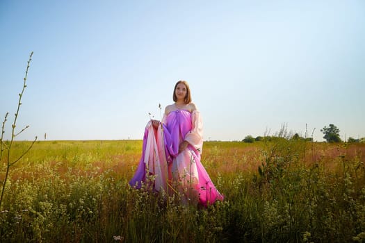 A woman wearing a colorful flowing garment enjoys the warm sunlight in a lush green field, surrounded by wildflowers and clear skies.