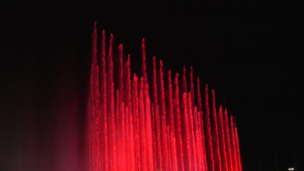 Nighttime view of numerous tall, slender red water jets forming a vibrant, vertical fountain display against a dark background.