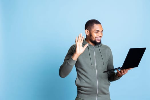 African american man chatting with his friends during videoconference call on laptop, stands against blue background. Young adult waving at videocall webcam, remotely meeting his mates.