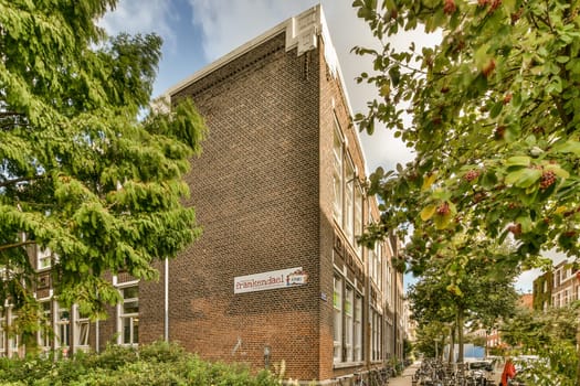 a brick building with bicycles parked on the sidewalk next to trees and bushes in front of it, under a cloudy blue sky