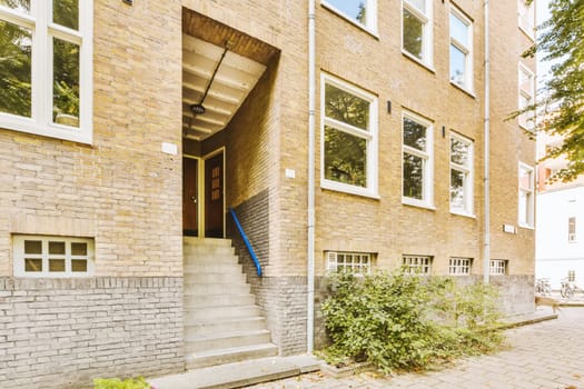 a brick building with some plants growing in the front and stairs leading up to the second floor, on a sunny day