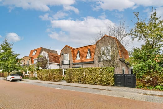 some houses on the side of a street with cars parked in front of them and green bushes growing along the road