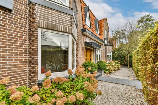 a brick house with flowers in the front and windows on either side, there is a gravel path leading up to it