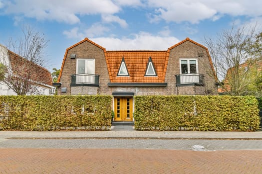 a brick house with a yellow front door and green hedges on either side of the house there is a blue sky in the background