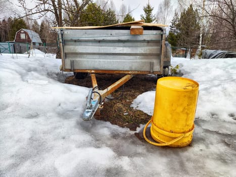 A metal trailer rests in a snowy area with a bright yellow barrel beside it, surrounded by melting snow and trees.