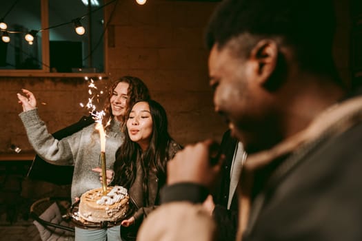 A group of friends joyfully celebrate a birthday outdoors in the evening. One friend holds a cake with sparklers while others cheer and smile, creating a festive atmosphere.