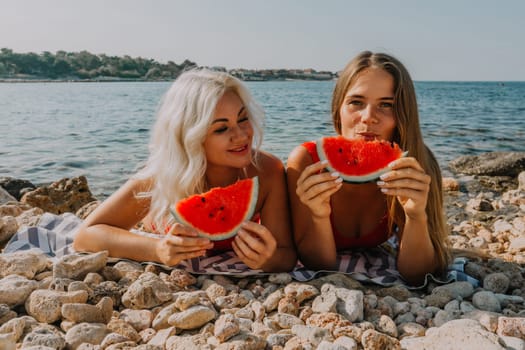 Two women lie on a pebbled beach, enjoying slices of watermelon by the sea. Sunny summer day.