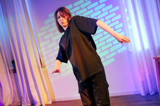 A dancer dressed in black performs fluid movements in a softly lit studio adorned with colorful light projections. The atmosphere is expressive and artistic, highlighting creativity.