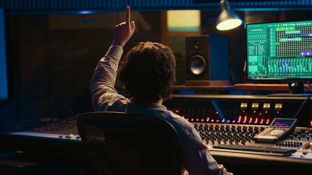 Sound engineer counting down for artist in vocal booth to start singing and producing new music, control room technical gear. Skilled audio technician operating knobs and sliders. Camera A.