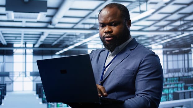 Computer scientist in high tech facility using laptop to patch network issues affecting server cabinets performance and connectivity, leading to poor client communication, camera B