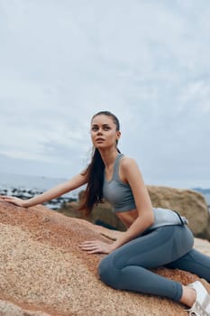 Fit young model in gray sportswear showcases strength and determination on a rocky beach, with a calm ocean backdrop and an inspiring attitude ideal for fitness themes