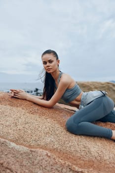 Fit young woman in gray sportswear poses on rocky beach, embodying strength and serenity, perfect for fitness, wellness, and outdoor activities themes