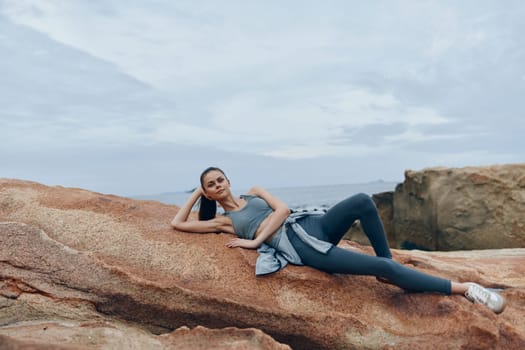 Fit female model in stylish sportswear lounges on coastal rocks, evoking relaxation and a healthy lifestyle against a tranquil ocean backdrop