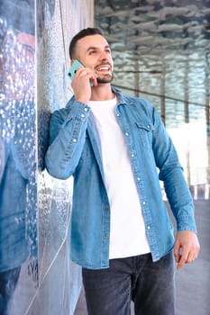 Close-up smiling man talking on a smartphone outdoors. He wears a denim jacket and stands near a reflective surface, enjoying a casual and modern communication moment in natural daylight. Vertical