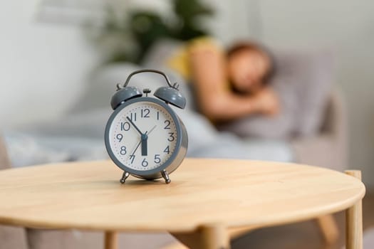 A classic alarm clock sits prominently, marking the beginning of a morning routine. The blurred figure of a woman in the background hints at her waking up.