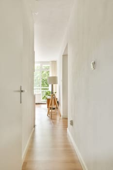 A bright and airy interior hallway featuring a minimalist design with wooden flooring and a modern lamp, inviting natural light from large windows.