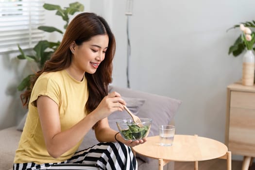 A young woman participates in her healthy morning routine, delighting in a bowl of fresh greens while seated comfortably in her stylish living space.